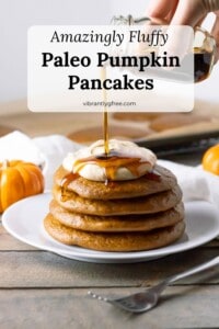 Side view of a stack of four fluffy paleo pumpkin pancakes topped with a swirl of pumpkin spice coconut whipped cream and a drizzle of maple syrup.