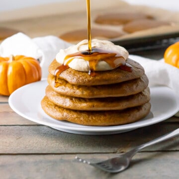 Side view of a stack of four fluffy paleo pumpkin pancakes topped with a swirl of pumpkin spice coconut whipped cream and a drizzle of maple syrup.