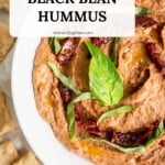 Overhead view of a serving bowl of black bean hummus garnished with sun-dried tomatoes and basil leaves.