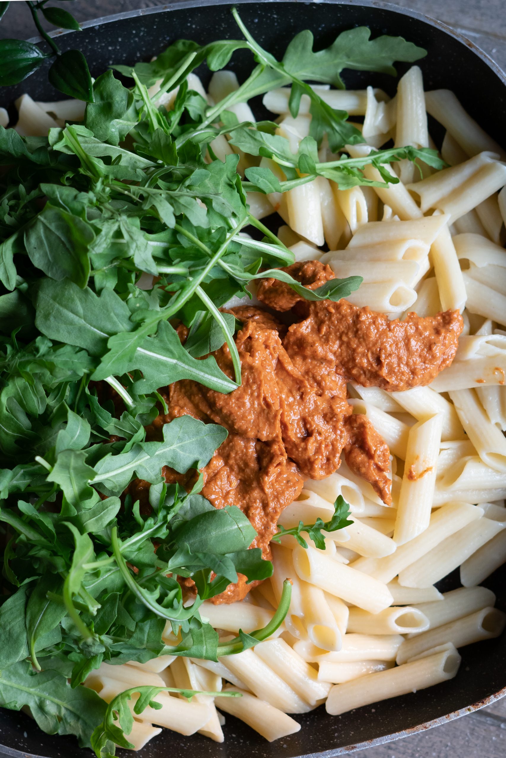 Fresh penne pasta, fresh arugula, and a freshly made batch of sauce in a pot, ready to be stirred together.