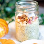 Side view of a glass jar of pumpkin spice chia pudding with apples and pumpkin seeds surrounded by mini pumpkins and green ivy.
