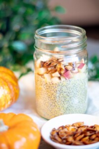 Side view of a glass jar of pumpkin spice chia pudding with apples and pumpkin seeds surrounded by mini pumpkins and green ivy.