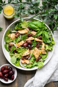 Tuscan Chicken Salad on a plate with fresh basil leaves, dressing in a jar, and kalamata olives.