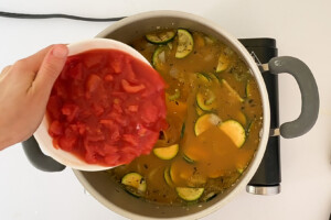 Overhead view of a hand adding canned diced tomatoes to the pot with veggies and broth.