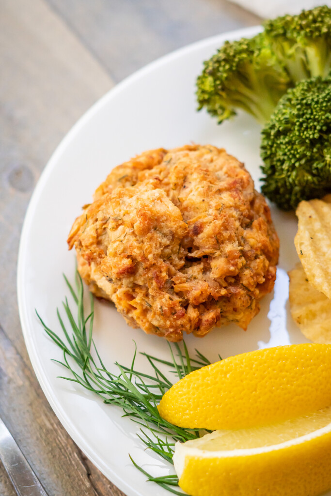 Side view of a baked salmon cake with steamed broccoli, dill pickle chips, lemon wedges and a sprig of fresh dill.
