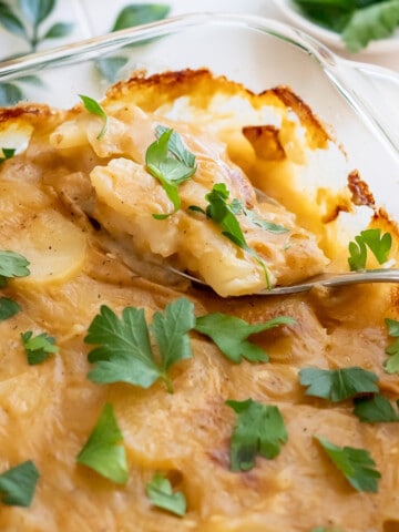 Side view of a glass dish of Dairy-Free Scalloped Potatoes with a spoon removing a bite to show the tender potatoes, smooth gravy, and oven-baked crust!