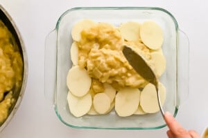 Overhead view of a layer of unevenly stacked potato slices with a hand spreading the first layer of sauce with a spoon.