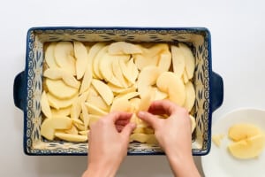 Overhead view of hands laying apple slices on top of the batter in a glass baking dish.