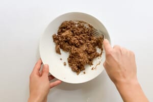Overhead view of hands mixing sugar, coconut oil, and gluten-free flour in a bowl with a fork.
