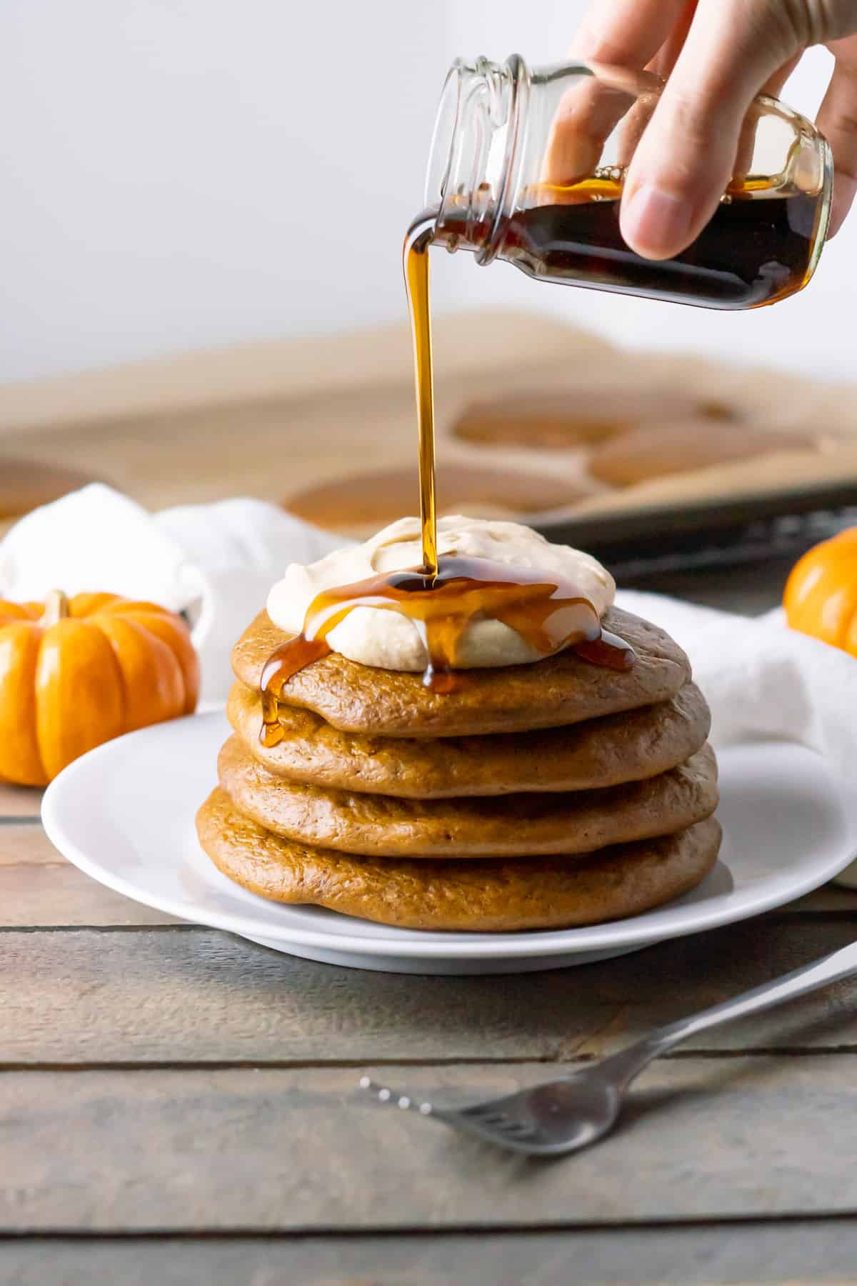 Side view of a tall stack of 4 Fluffy Paleo Pumpkin Pancakes with pumpkin spice whipped cream and a hand drizzling maple syrup from a jar!