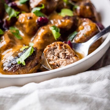 Side view of Turkey Cranberry Meatballs with smooth buttery gravy and garnished with dried cranberries and chopped parsley. One meatball is cut in half to show the texture and variety.
