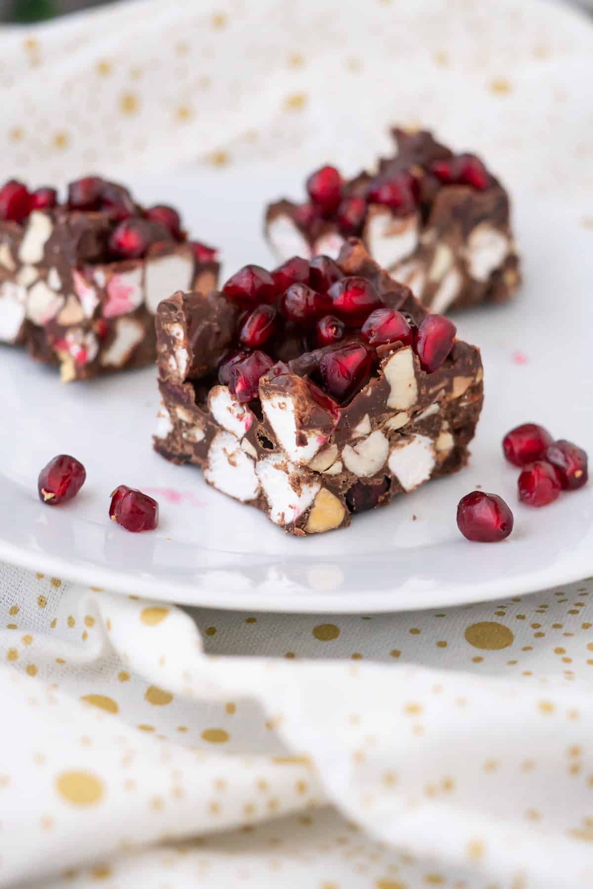 Side view of three squares of Christmas Rocky Road on a plate to show layers of chocolate, almonds, marshmallows, and pomegranate seeds.