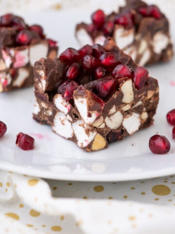 Side view of three squares of Christmas Rocky Road on a plate to show layers of chocolate, almonds, marshmallows, and pomegranate seeds.