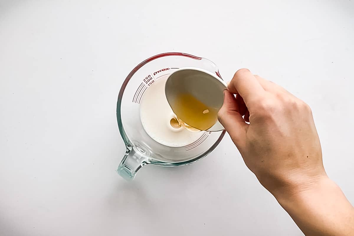 Pouring apple cider vinegar into a glass measuring cup with almond milk.