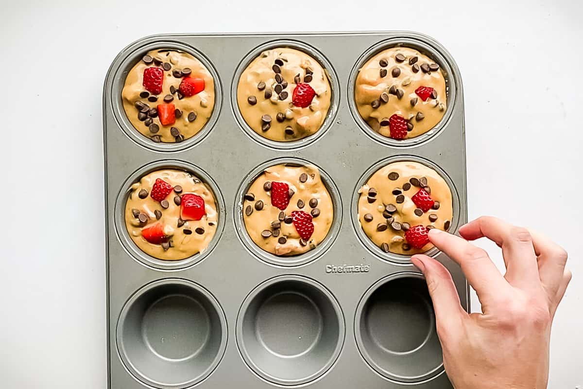 A hand adding chocolate chips and pieces of strawberry to the tops of the muffin batter just before baking for decoration.