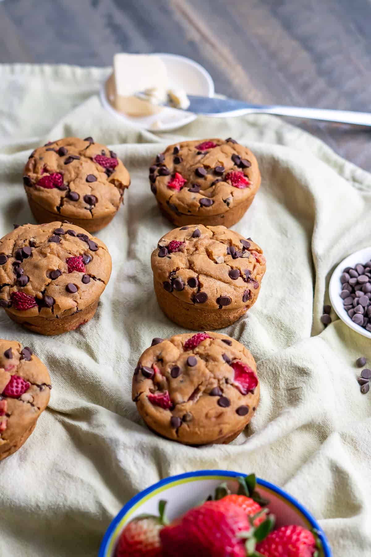 Six small batch gluten free strawberry muffins with chocolate chips sitting on a cloth napkin with butter and chocolate chips on the side.
