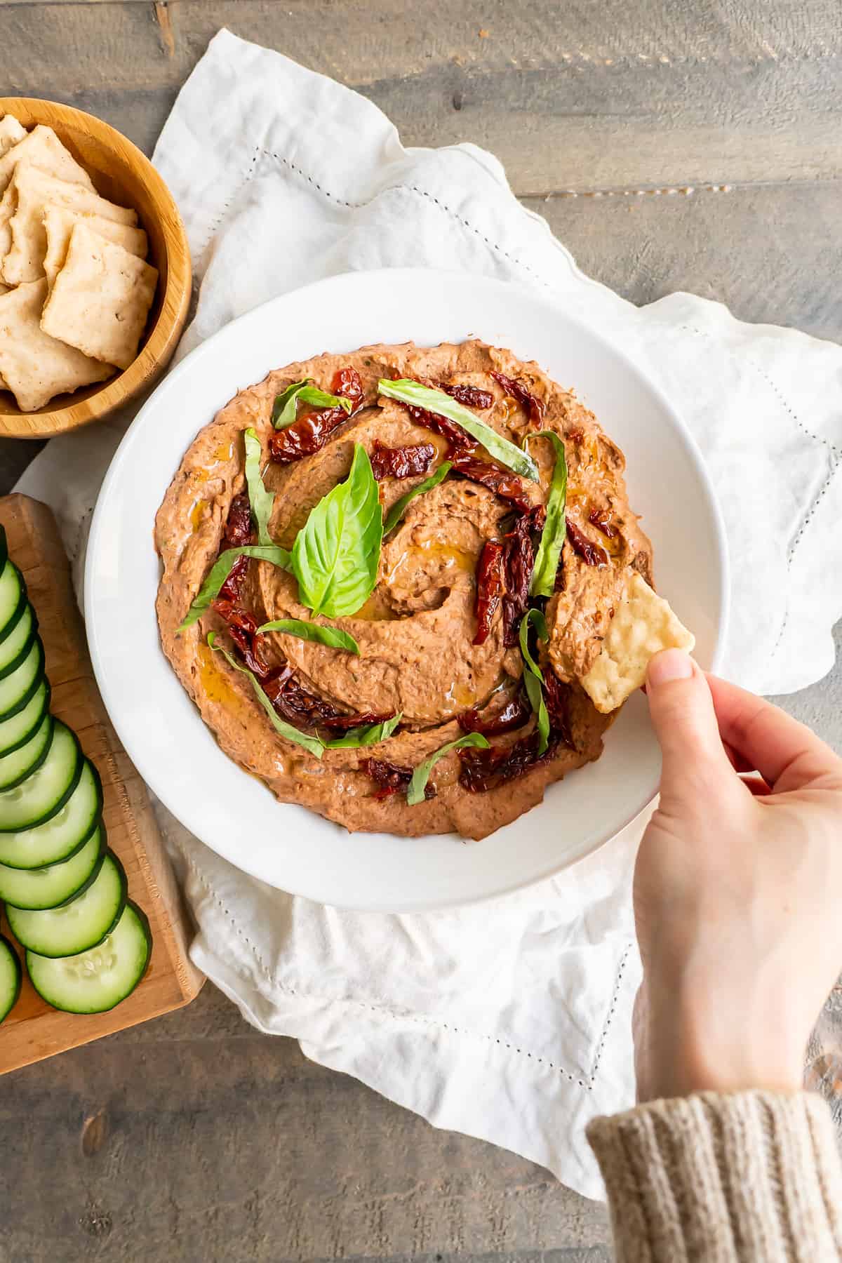 A serving bowl of creamy hummus garnished with sun-dried tomatoes and basil with gluten-free crackers.