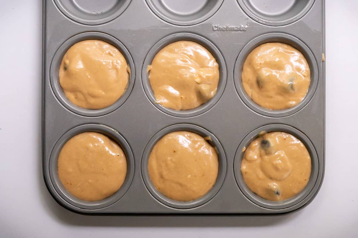 Six cups of a standard muffin tin filled with gluten free blueberry muffin batter.