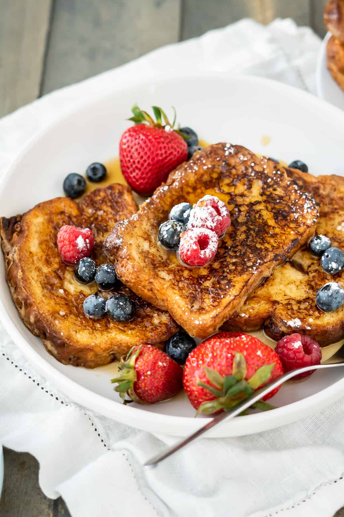 A stack of three pieces of french toast with fresh berries, powdered sugar, and maple syrup.