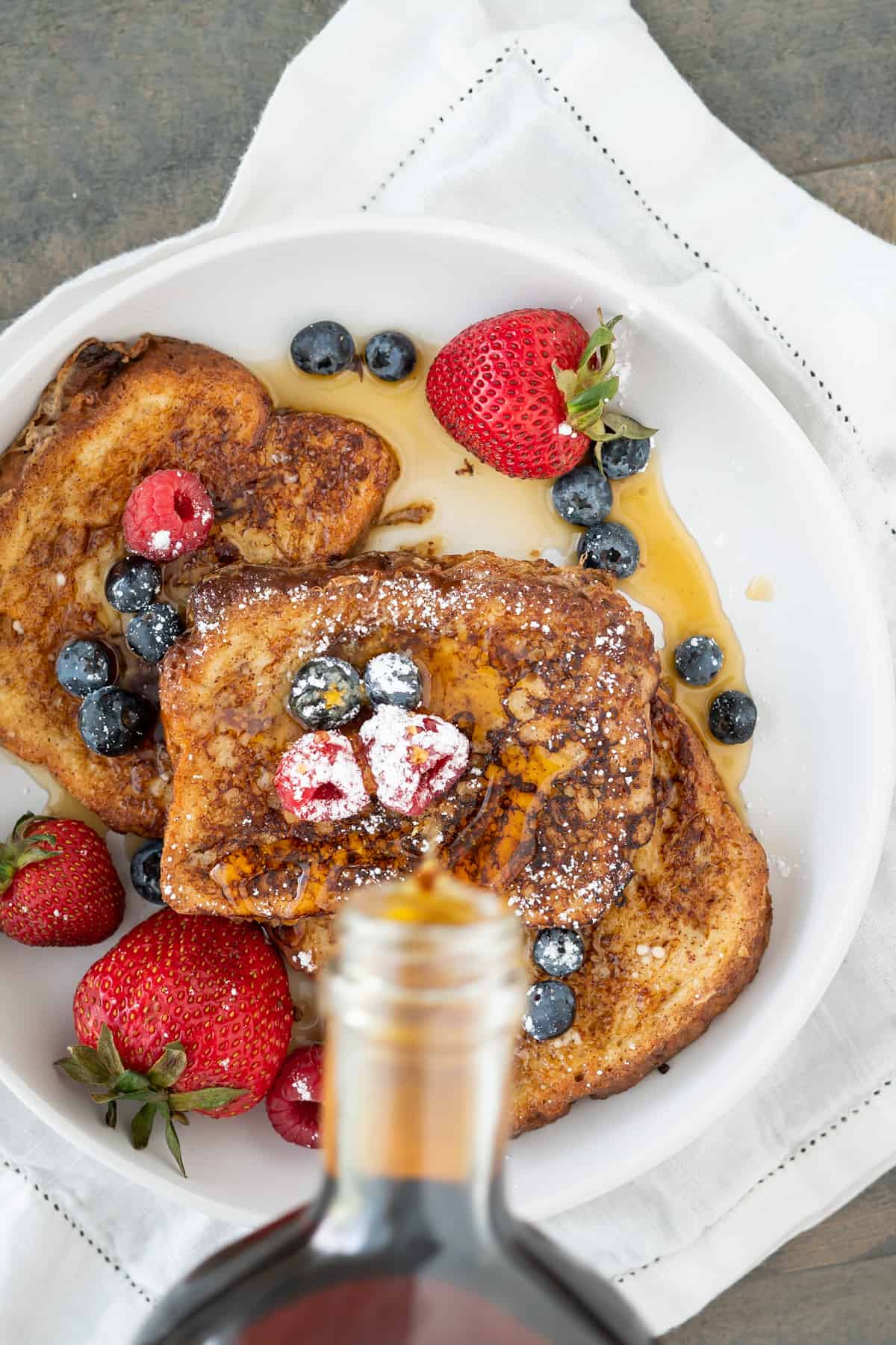Overhead view of pouring maple syrup over slices of dairy-free french toast with berries and powdered sugar.