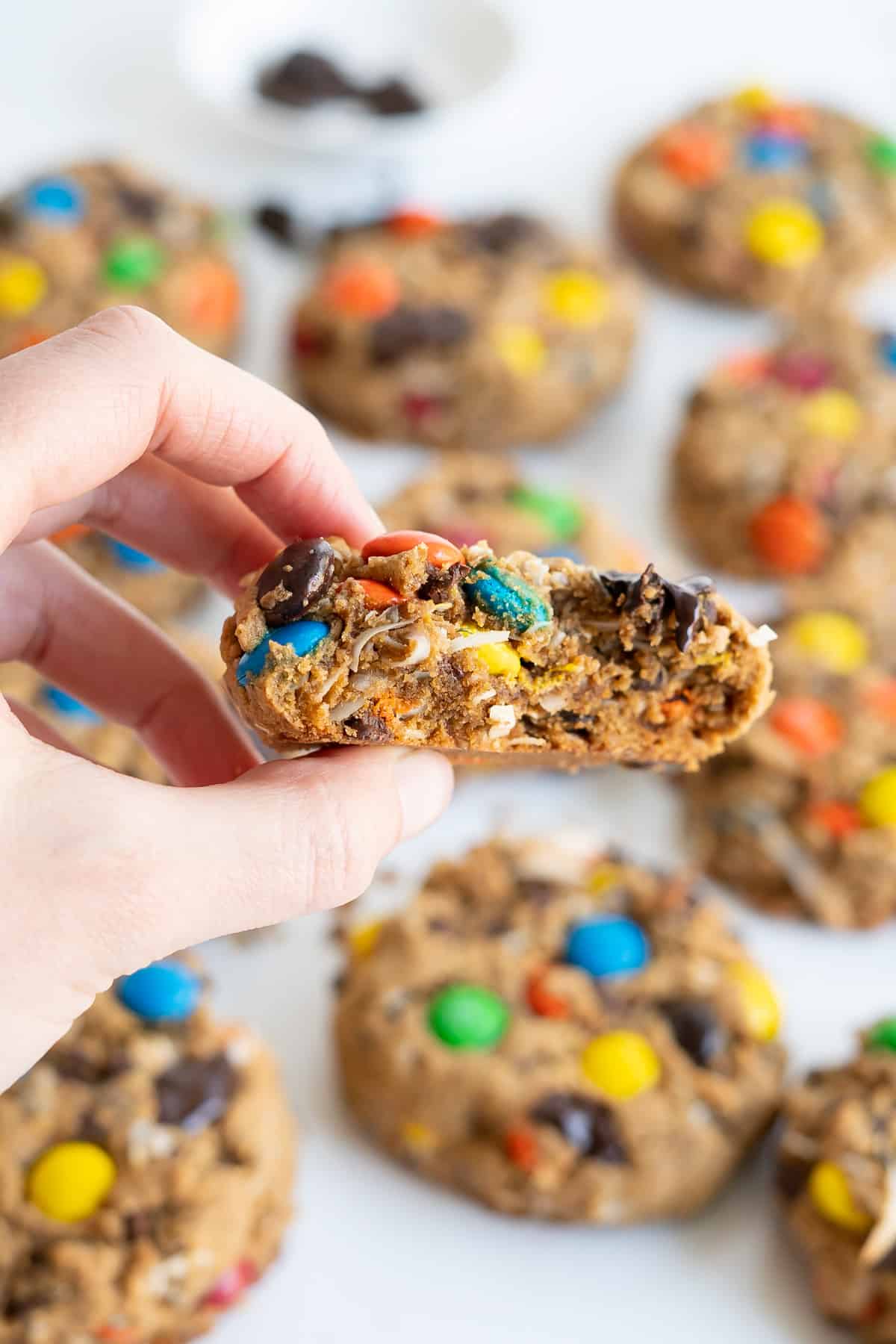 A hand holding half of a gluten-free monster cookie to show the chewy texture with M&M's, shredded coconut, and chocolate chips.