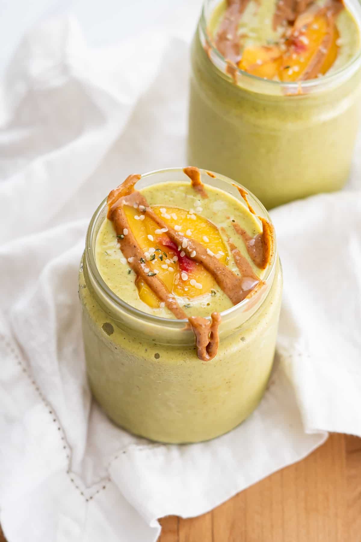 Side view of a jar with peach mango green smoothie topped with peach slices, hemp seeds, and almond butter.