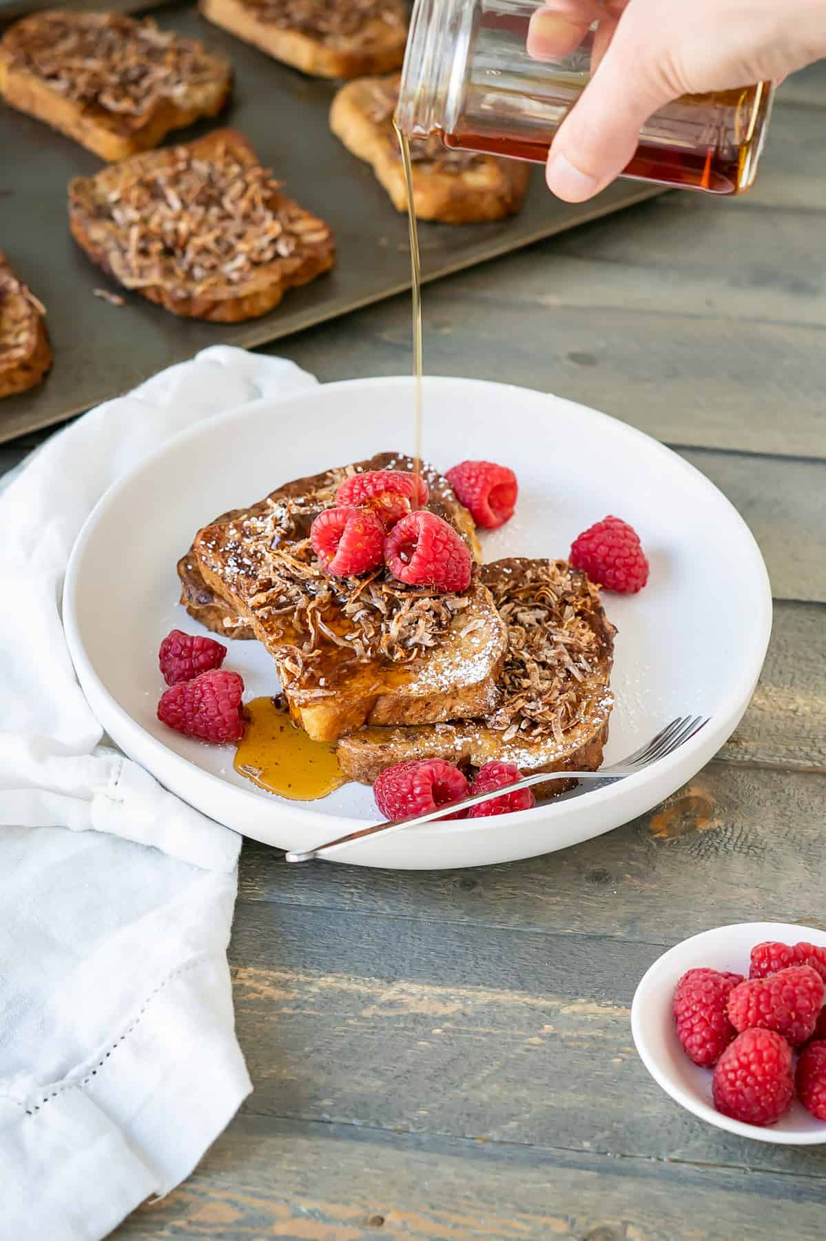 A hand pouring syrup over a coconut french toast stack with fresh raspberries.