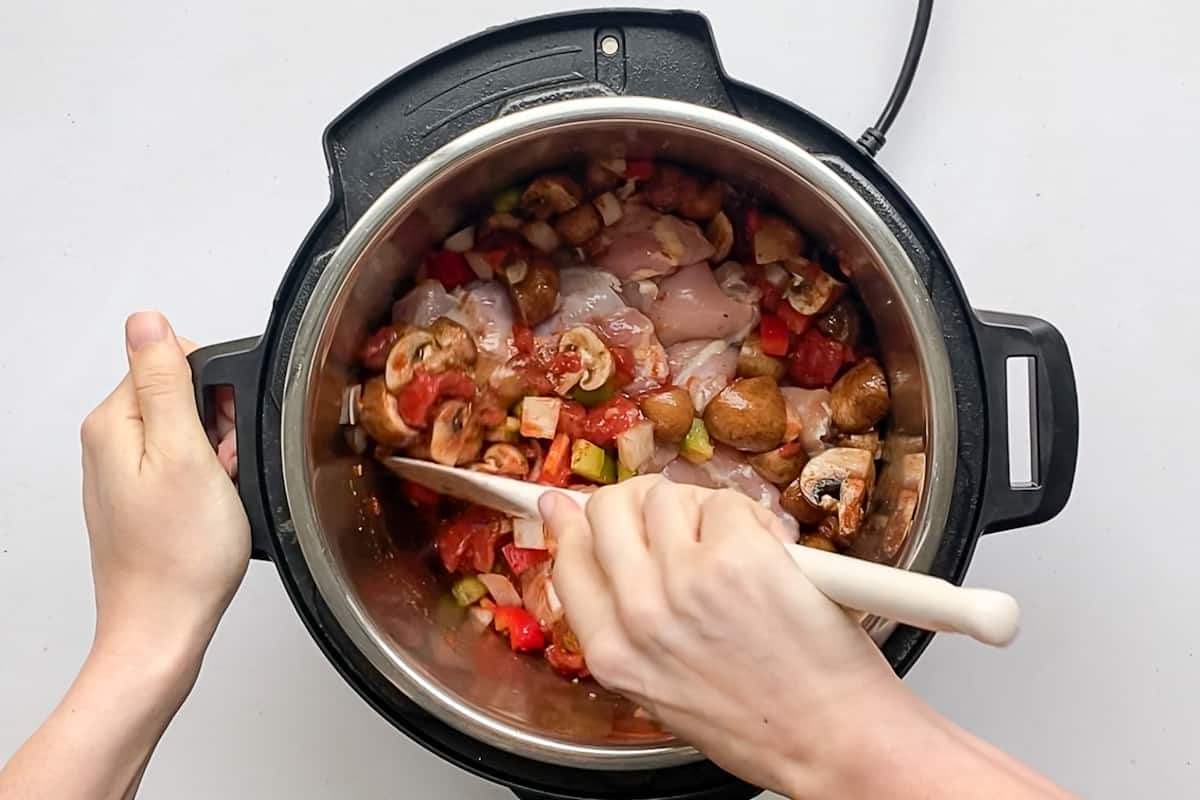 A hand using a spatula to push vegetables over top of chicken thighs nestled in an instant pot.
