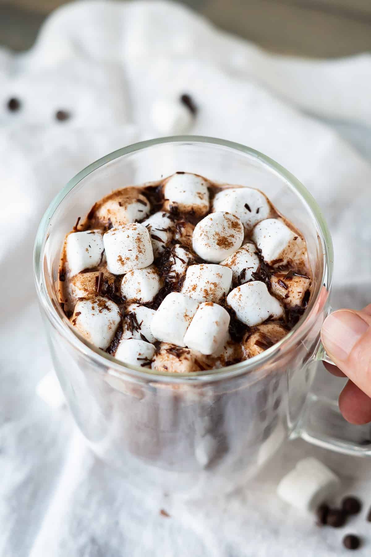 A glass mug full of hot chocolate topped with marshmallows, a sprinkle of cinnamon, and chocolate shavings.