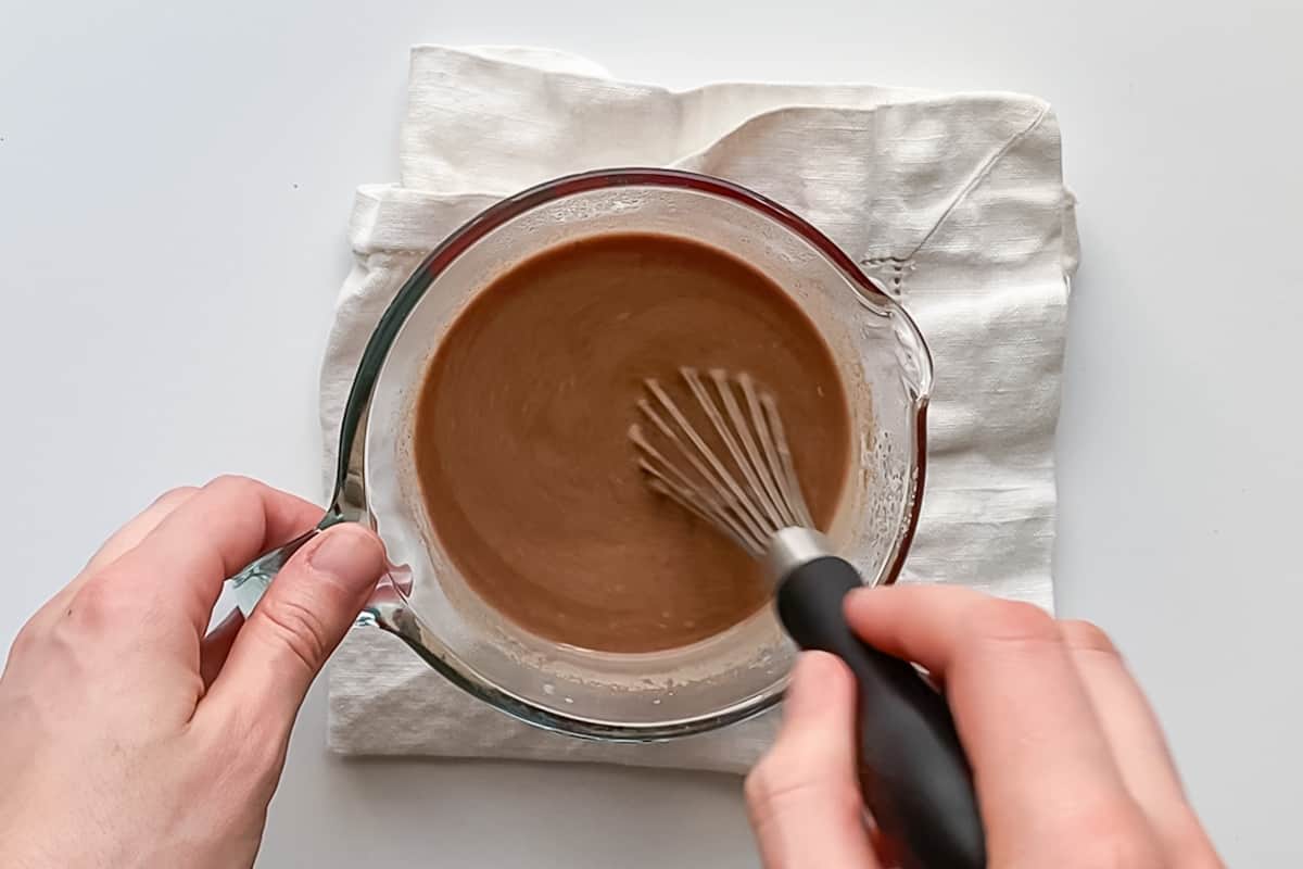 Hands holding a whisk and mixing hot chocolate in a liquid measuring cup.