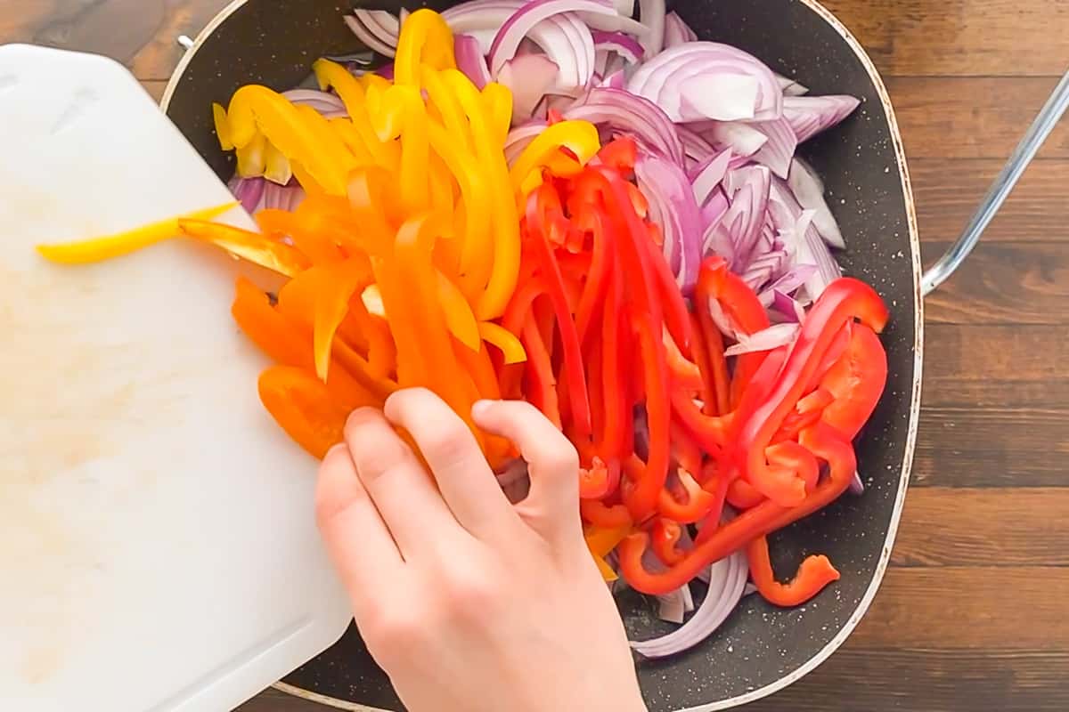 A hand sliding a pile of sliced red onions and colorful bell peppers into a large skillet.