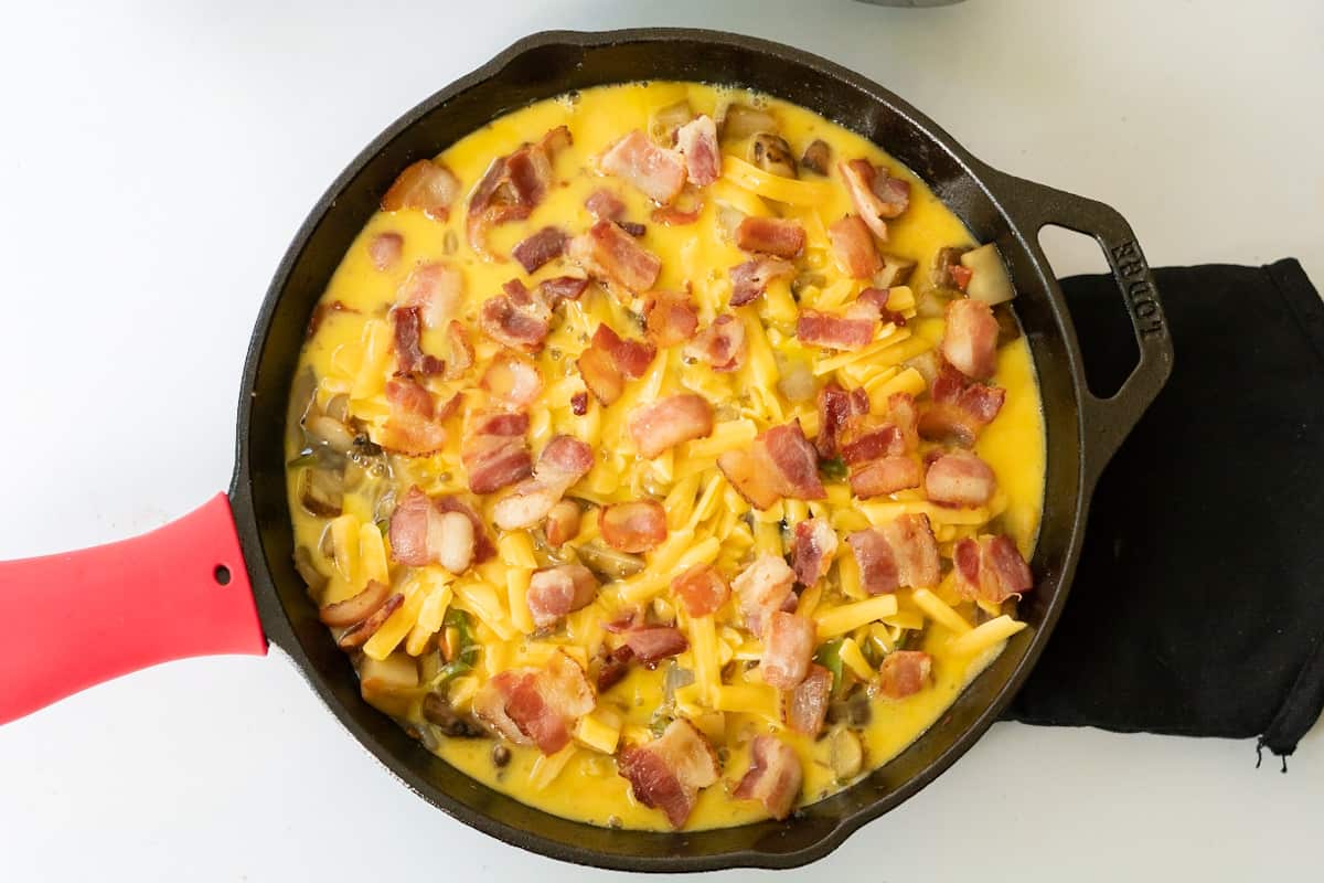 A cast iron skillet before baking with crisp bacon, wet egg, and unmelted cheese.