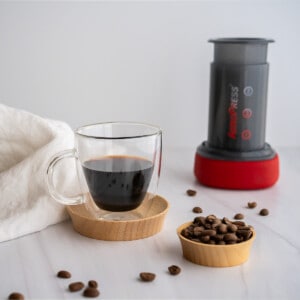 A clear glass espresso cup with two shots or aeropress espresso surrounded by an aeropress and espresso beans.