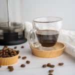 A double shot of french press espresso in a clear glass espresso cup with the french press and espresso beans beside it.