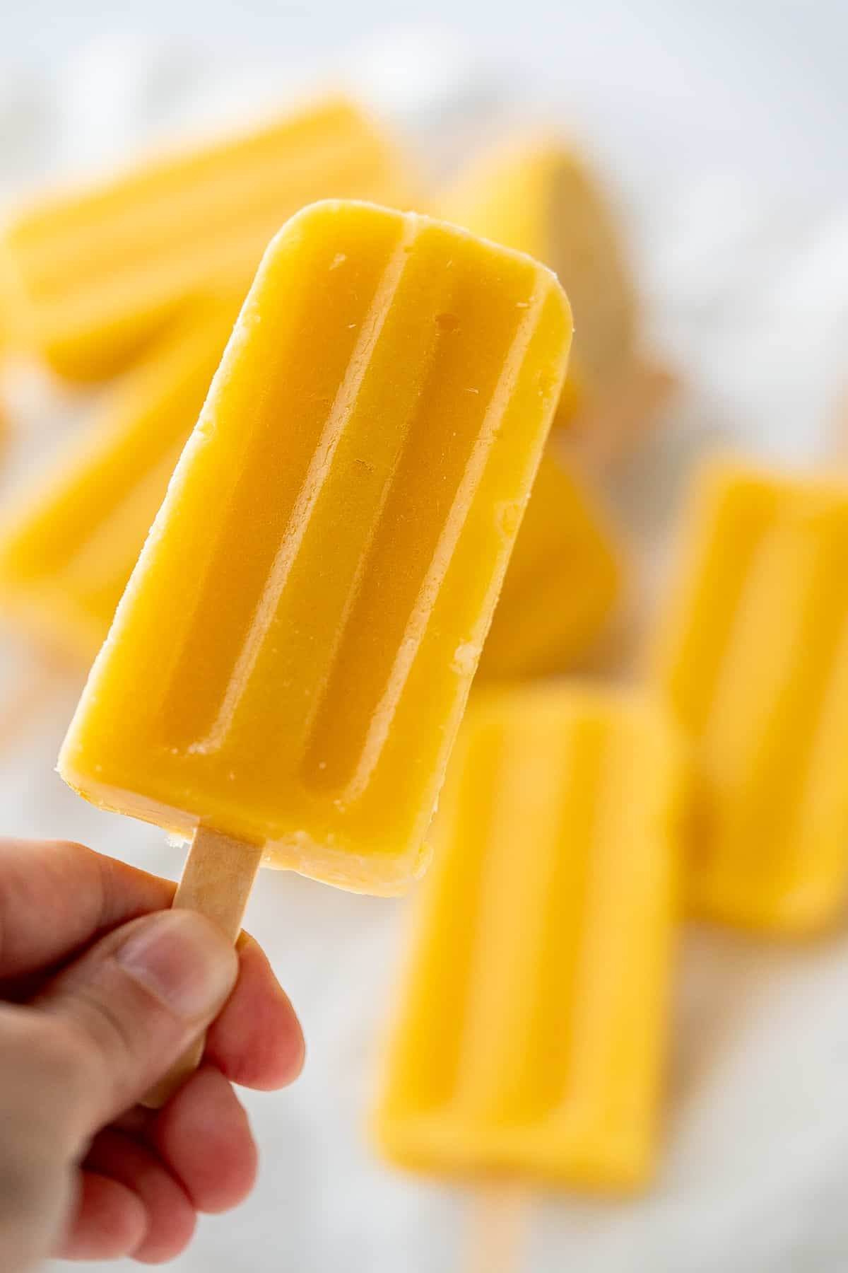 A hand holding the stick of a healthy mango popsicle to show it's soft texture and vibrant mango color.