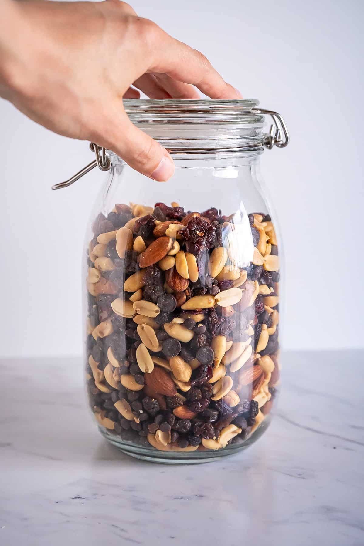 A tall storage jar filled with trail mix that includes peanuts, almonds, dried fruit, and chocolate chips.