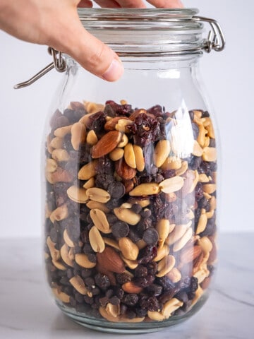 A tall storage jar filled with trail mix that includes peanuts, almonds, dried fruit, and chocolate chips.