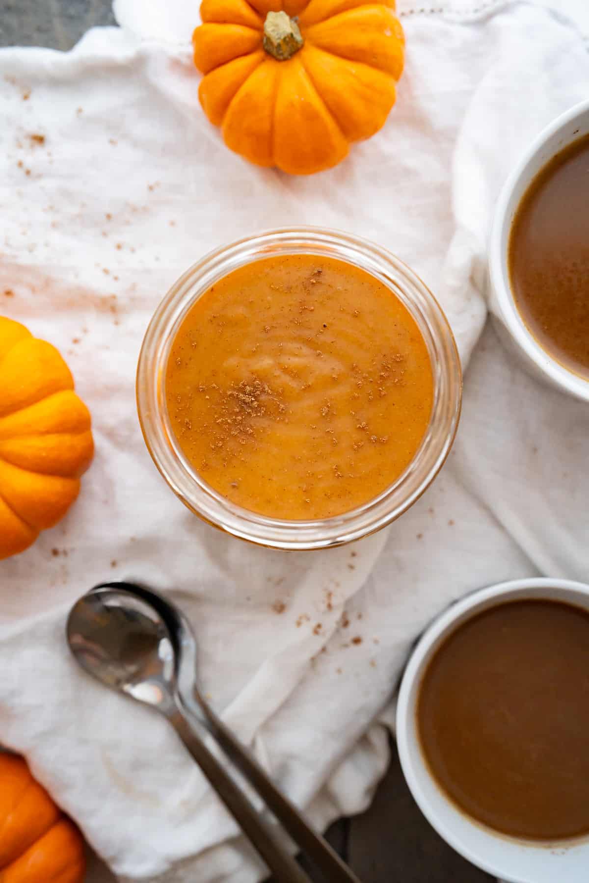 A jar of homemade pumpkin spice creamer surrounded by two pumpkin coffees and mini pumpkins.