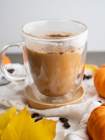 A homemade pumpkin spice latte in a clear mug surrounded by fall leaves and mini pumpkins.