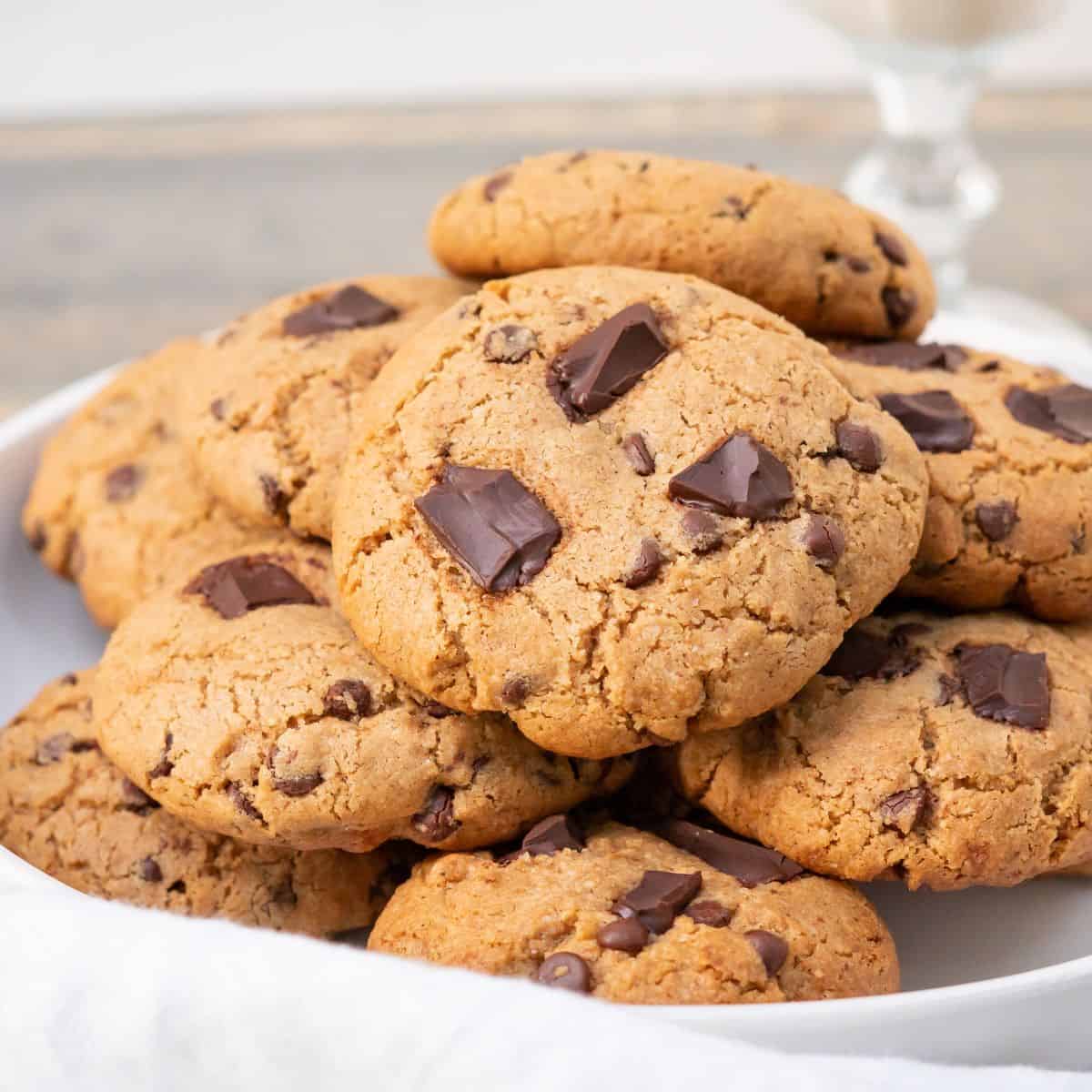 A pile of freshly baked golden brown almond flour chocolate chip cookies with melted chocolate chunks on top and a tall glass of milk.