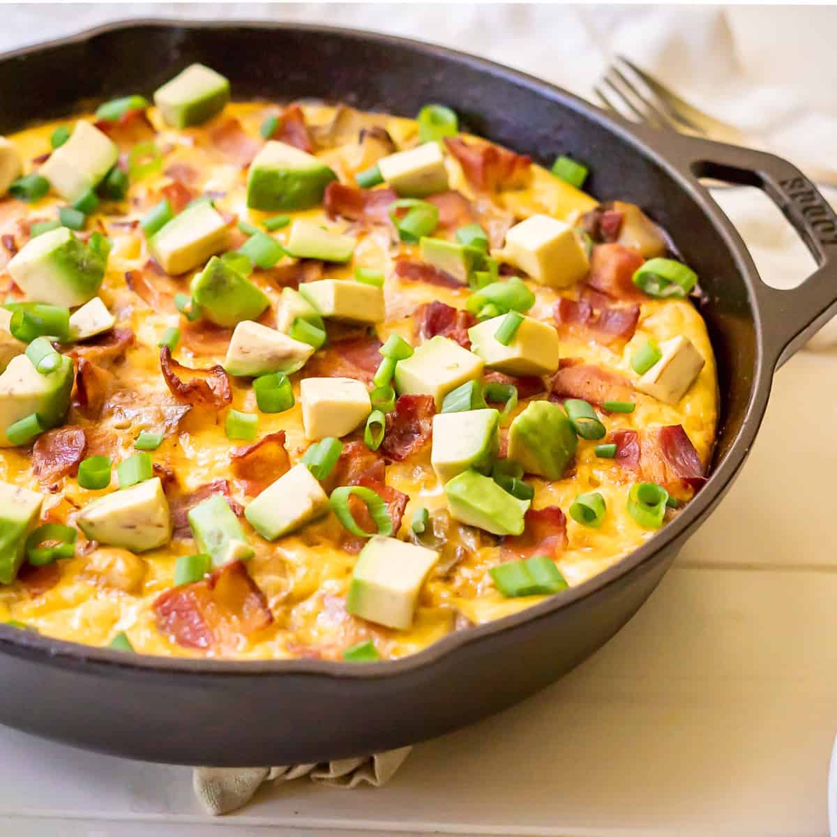 A cast iron skillet filled with a baked Southwest breakfast skillet topped with chopped avocado, bacon, and green onions.