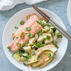 A salmon sushi bowl with cooked salmon, cucumber, avocado, green onions, rice and a sriracha mayo sauce.