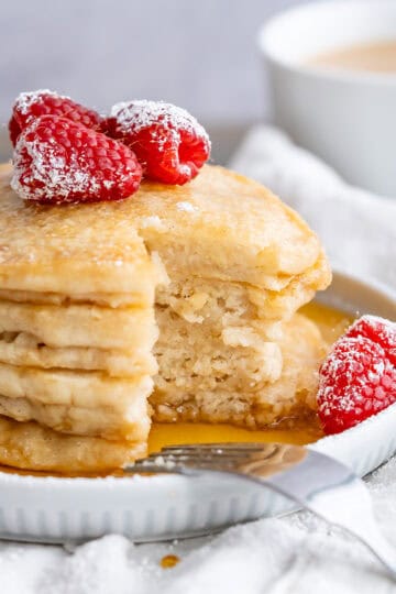 A stack of four fluffy gluten-free pancakes with maple syrup and fresh raspberries.