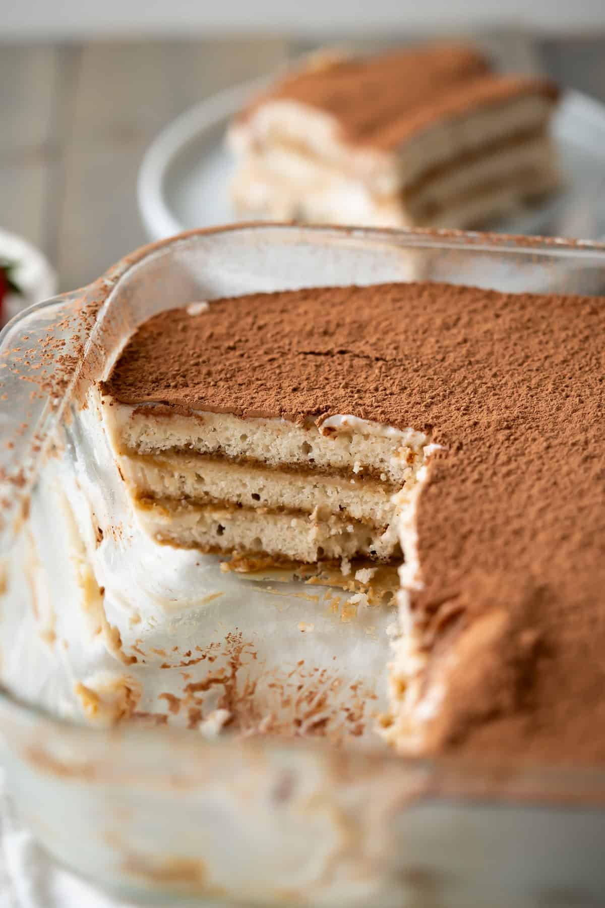 A piece removed from a glass dish of vegan tiramisu to show the layers of vegan ladyfingers, custard, and cocoa powder.