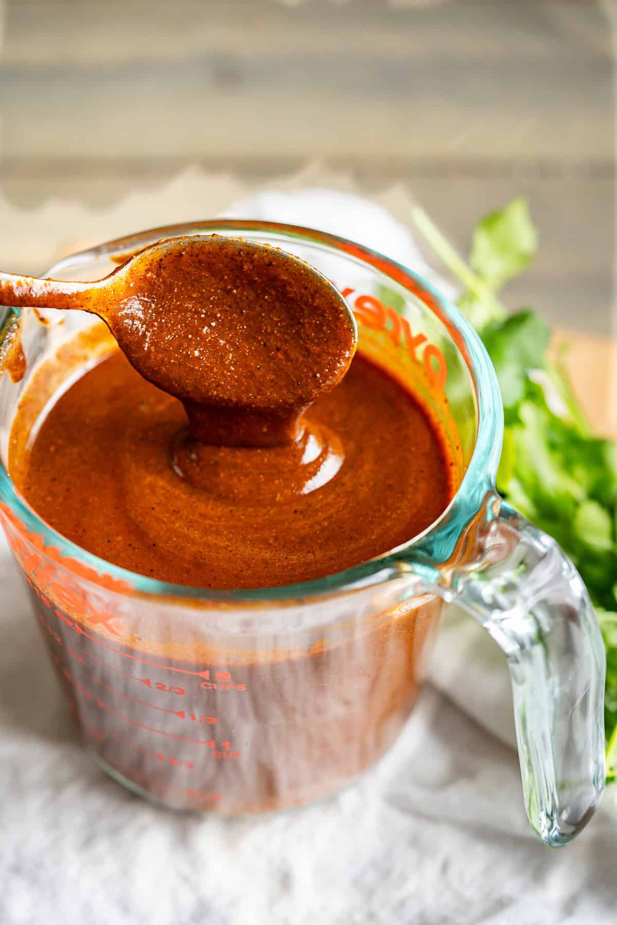 A spoon dripping homemade enchilada sauce into a liquid measuring cup to show the thickness.