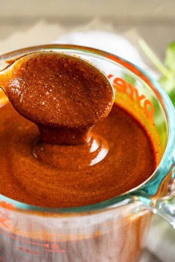 A spoon dripping homemade enchilada sauce into a liquid measuring cup to show the thickness.