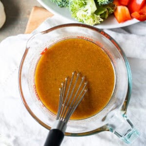 A stir-fry sauce with whisk beside a plate of stir-fry vegetables