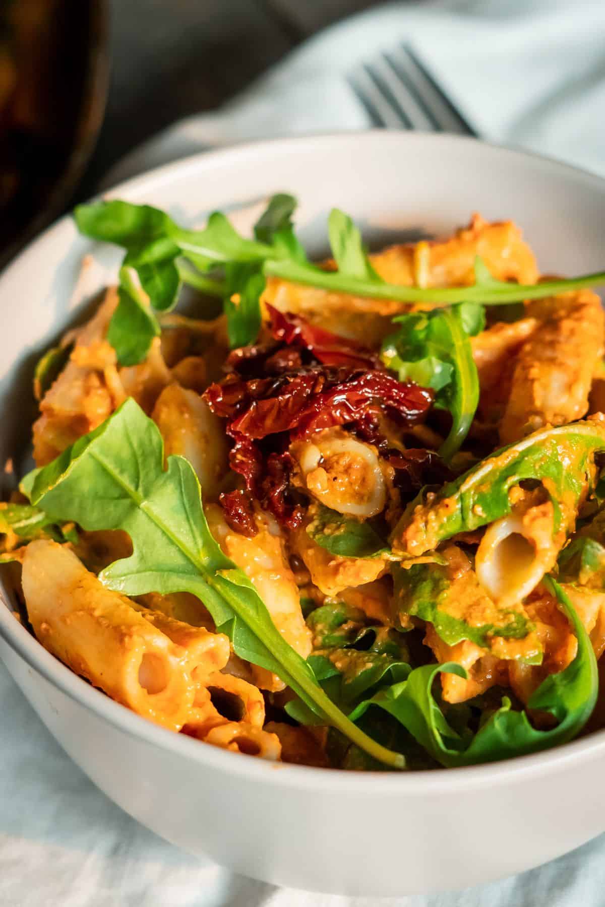 A bowl of pasta with a creamy red sauce, topped with sun-dried tomatoes and arugula.