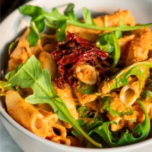 A bowl of pasta with a creamy red sauce, topped with sun-dried tomatoes and arugula.