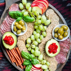 A gluten and dairy-free charcuterie board with fruits, olives, avocado, roasted red peppers, meats, and rice crackers.
